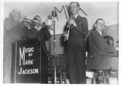 "MUSIC BY MARK JACKSON" IS WHAT IS PRINTED ON THE PODIUM, THREE MEN STANDING PLAYING BRASS INSTRUMENTS AND ONE MAN SITTING PLAYING PIANO